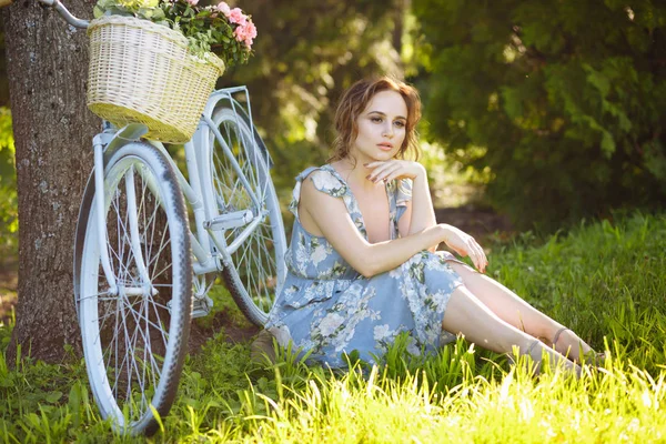 Porträt eines schönen Mädchens im Wald, auf dem Gras sitzend, neben dem Fahrrad, mit einem Blumenkorb, hinter den Sonnenstrahlen, einem blau geblümten Kleid, Sommerspaziergang. — Stockfoto