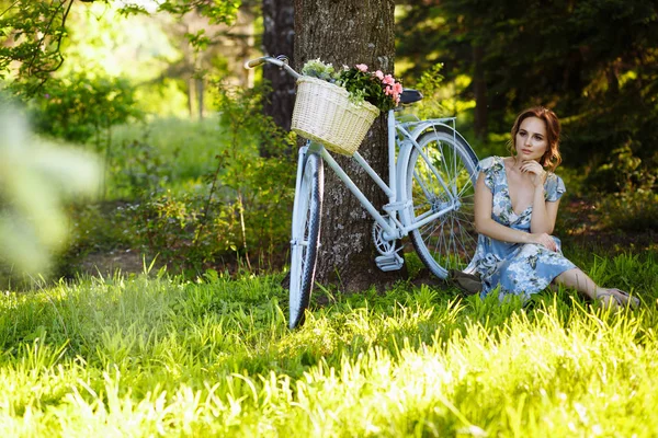 Retrato de uma menina bonita na floresta, sentado na grama, ao lado da bicicleta, com uma cesta de flores, atrás dos raios do sol, um vestido de flor azul, passeio de verão . — Fotografia de Stock