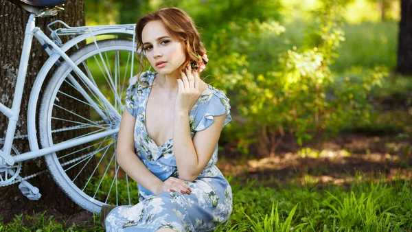 Portrait of a beautiful girl in the forest, sitting on the grass, next to the bike, with a basket of flowers, behind the rays of the sun, a blue flowered dress, summer walk. — Stock Photo, Image