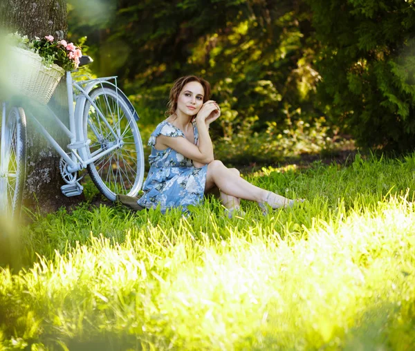 Retrato de una hermosa niña en el bosque, sentada en la hierba, junto a la bicicleta, con una cesta de flores, detrás de los rayos del sol, un vestido de flores azules, paseo de verano . — Foto de Stock