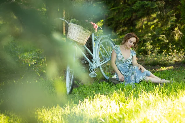 Portrait d'une belle fille dans la forêt, assise sur l'herbe, à côté du vélo, avec un panier de fleurs, derrière les rayons du soleil, une robe à fleurs bleues, promenade estivale . — Photo