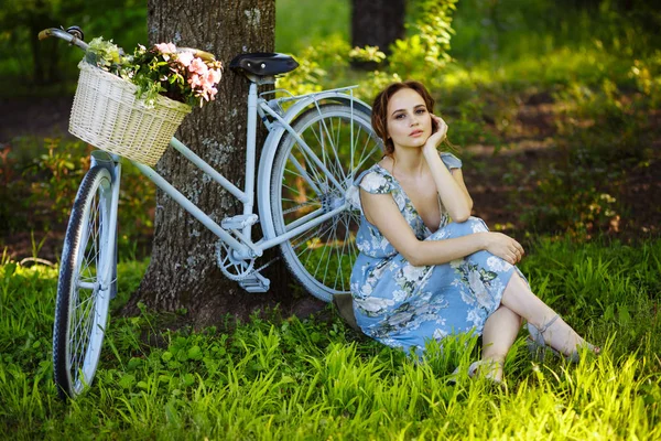 Portret pięknej dziewczyny w lesie, siedząc na trawie, obok roweru, z koszem kwiatów, za promieniami słońca, niebieską kwiecistką sukienkę, letni spacer. — Zdjęcie stockowe
