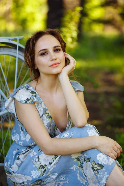 Ormanda güzel bir kız portresi, çim üzerinde oturan, bisiklet yanında, çiçek sepeti ile, güneş ışınları arkasında, mavi çiçekli elbise, yaz yürüyüşü. — Stok fotoğraf