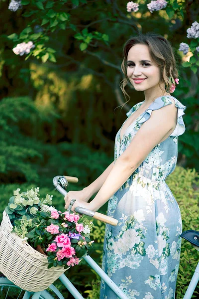 Retrato de uma menina bonita na floresta, segurando uma bicicleta com uma cesta de flores, atrás dos raios do sol, um vestido de flor azul, passeio de verão — Fotografia de Stock