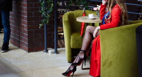 Дівчина в червоному плащі в ажурних колготках, сидить в кафе над чашкою кави, поруч з нею червона парасолька чекає на зустріч — стокове фото