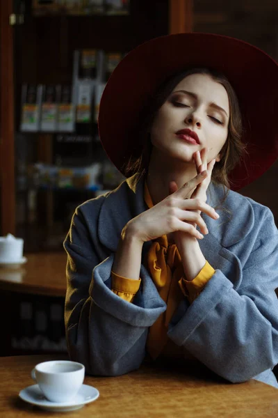Chica en un café con una taza de café y un hat.portrait de la joven sensual con sombrero floppy y blusa con lazo. Hermosa mujer morena en el café sosteniendo la taza de café — Foto de Stock