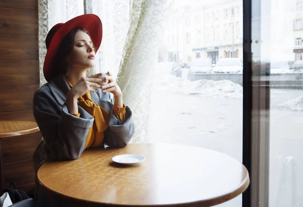 Ragazza in un caffè con una tazza di caffè e un cappello.ritratto di ragazza sensuale che indossa cappello floppy e camicetta con fiocco. Bella donna bruna in caffè che tiene una tazza di caffè — Foto Stock