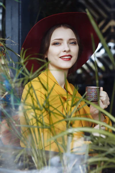 Chica en un café con una taza de café y un hat.portrait de la joven sensual con sombrero floppy y blusa con lazo. Hermosa mujer morena en el café sosteniendo la taza de café — Foto de Stock