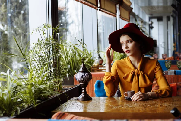 Chica en un café con una taza de café y un hat.portrait de la joven sensual con sombrero floppy y blusa con lazo. Hermosa mujer morena en el café sosteniendo la taza de café —  Fotos de Stock