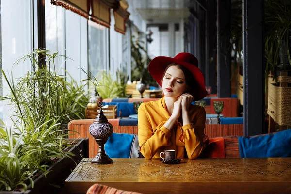Jente på en kafe med en kopp kaffe og et hat. portrett av en sensuell ung jente med slapp hatt og bluse med bue. Vakker brunettdame på kafeen med kaffekopp – stockfoto
