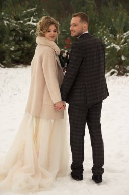 Çift sarılıyor ve öpücüğe yakın, el ele tutuşuyor, ellerinde bir buket çiçek. Kelebekli bir takım elbise ve elbiseli bir ceket. Gelin ve damat