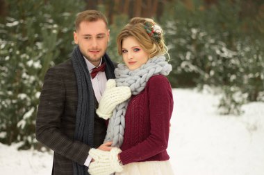 Çift sarılıyor ve öpücüğe yakın, el ele tutuşuyor, ellerinde bir buket çiçek. Kelebekli bir takım elbise ve elbiseli bir ceket. Gelin ve damat