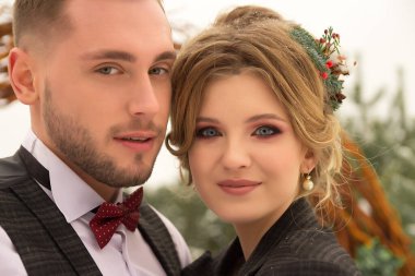 Çift sarılıyor ve öpücüğe yakın, el ele tutuşuyor, ellerinde bir buket çiçek. Kelebekli bir takım elbise ve elbiseli bir ceket. Gelin ve damat