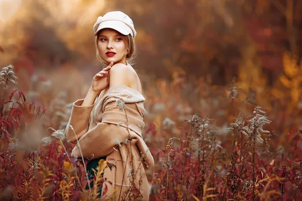 Hermosa chica en una gorra y un abrigo con un hombro desnudo en el fondo del otoño — Foto de Stock