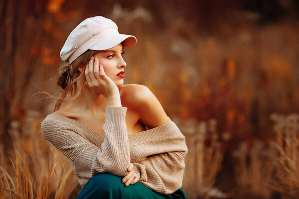 Beautiful girl in a cap and coat with a bare shoulder on the background of autumn walk green skirt, sitting