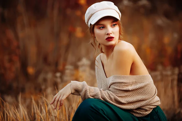 Hermosa chica en una gorra y un abrigo con un hombro desnudo en el fondo del otoño — Foto de Stock