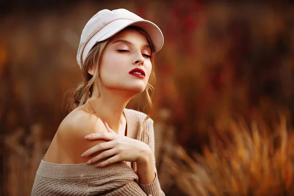 Hermosa chica en una gorra y un abrigo con un hombro desnudo en el fondo del otoño — Foto de Stock