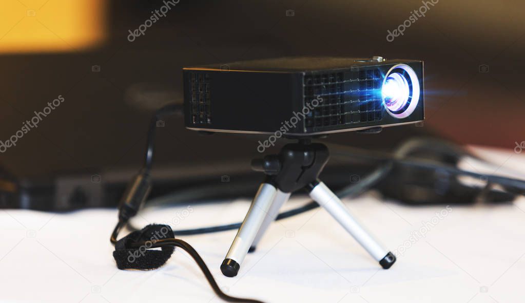 Black projector with tripod installed on white table, in hall or seminar meeting room. Real projector at business conference or presentation in the office room. Close up of projector for cinema and video projections. Business and education concept.