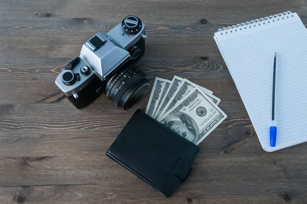 A retro camera, a purse with money and a notebook with a pen on a wooden table.