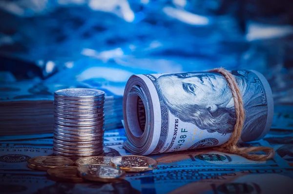 A roll of dollars with coins on the background of scattered one hundred dollar bills in blue light