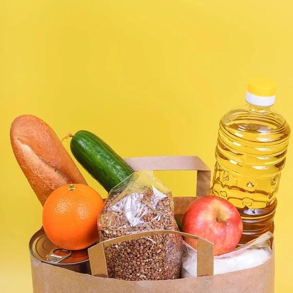 Paper bag with food supplies for the period of quarantine isolation on a yellow background. Delivery, donation, coronavirus. Copyspace. Buckwheat pasta sugar peas canned food tomatoes cucumber bread orange apples eggs ginger.