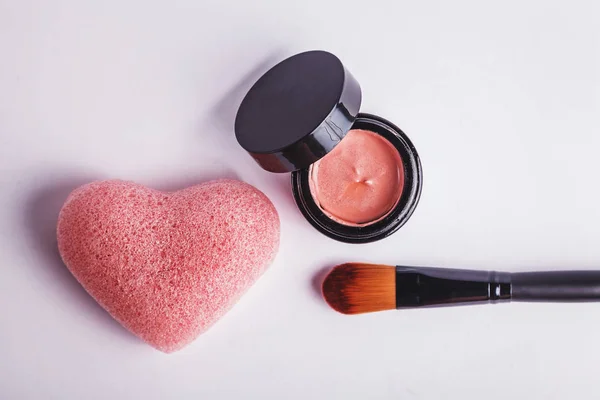 Cosmetic set of pink clay face mask, brush and light pink heart shaped konjac sponge. Top view on white background.
