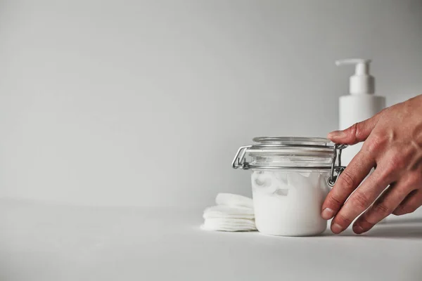 一个女人的手触摸一个透明的玻璃瓶充满了白色的身体奶油在白色背景 — 图库照片