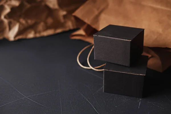 Two matte black carton gift boxes laying on a black painted scratched surface, craft paper bag blurred on background, home gift wrapping craft theme
