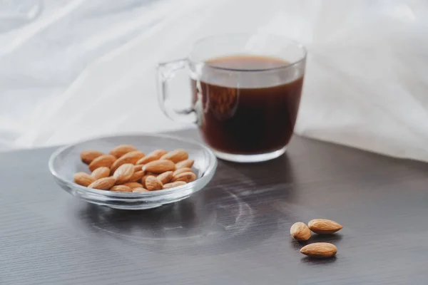 Concept of helathy snack - almonds with coffee placed on a dark laminate wood surface with white curtain on background on a very sunny day