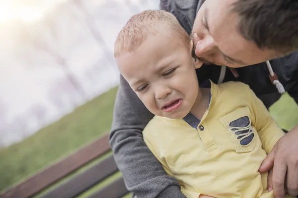 Closeup portrait of a young father and crying kid on playground. Young father hugs crying child. Dad and son.