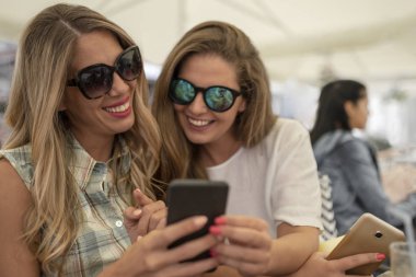 Seyir telefon kafede otururken iki neşeli neşeli kız. Mutlu genç kadın kahve dükkanında eğleniyor. Arkadaşlarım serbest zaman zevk.