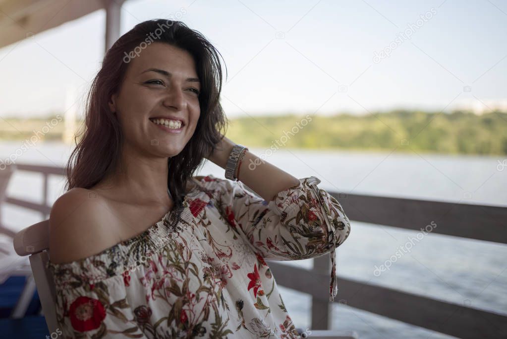 Portrait of beautiful young girl on sunny day in nature, near river. Cheerful woman model sitting in cafe enjoying free time. Pretty girl having fun outdoor.