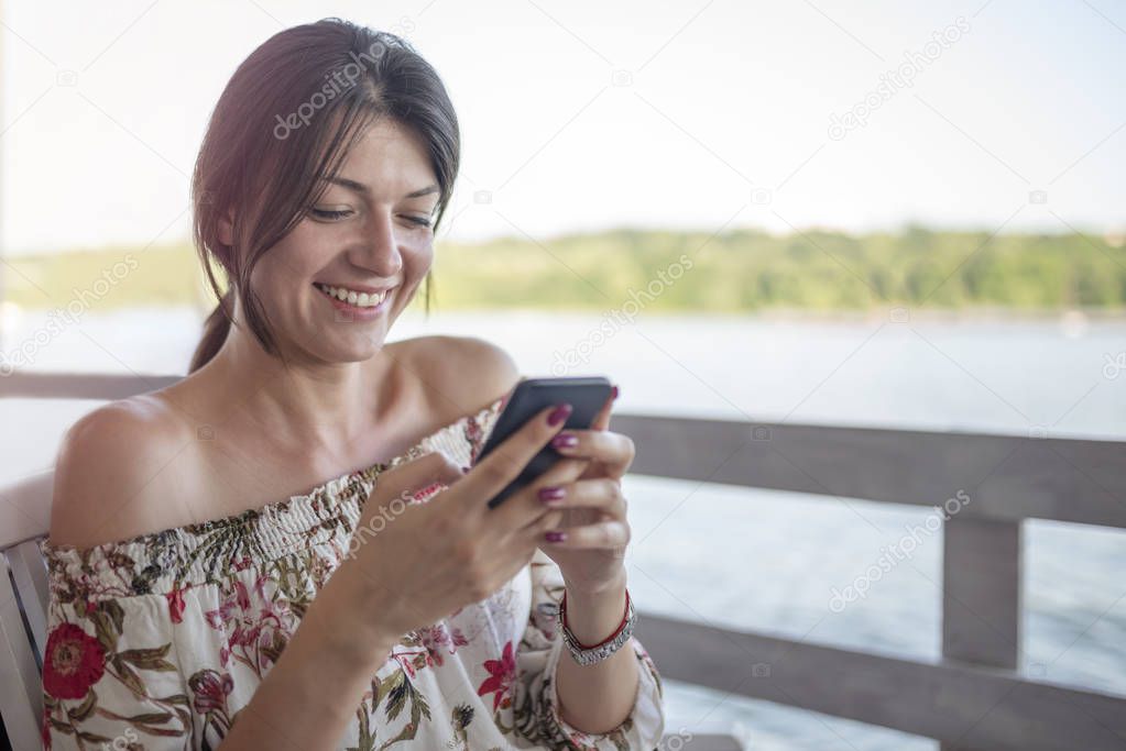 Portrait of beautiful young girl with phone on sunny day in nature, near river. Cheerful woman model sitting in cafe enjoying free time. Pretty girl talking on mobile phone.