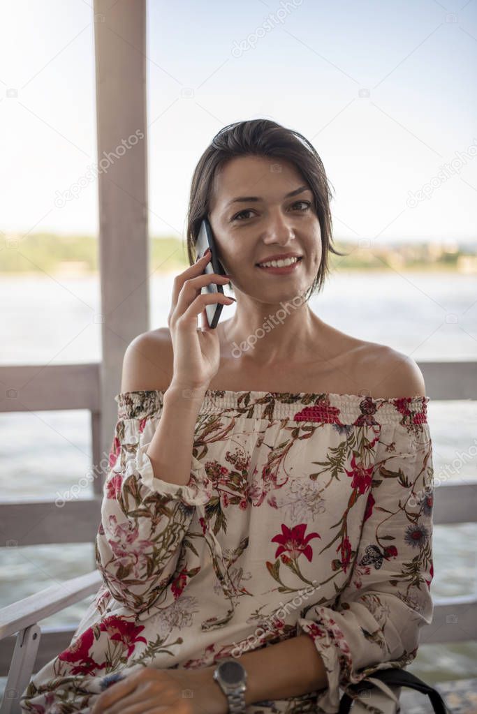 Portrait of beautiful young girl with phone on sunny day in nature, near river. Cheerful woman model sitting in cafe enjoying free time. Pretty girl talking on mobile phone.