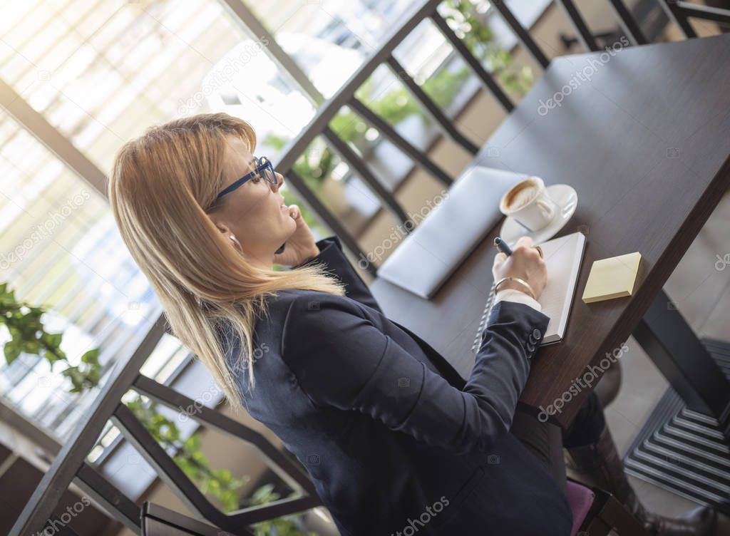 Business woman taking notes and drinkinkg coffee in restaurant. Blonde girl solving business problems in coffee shop. Woman in blue suit.