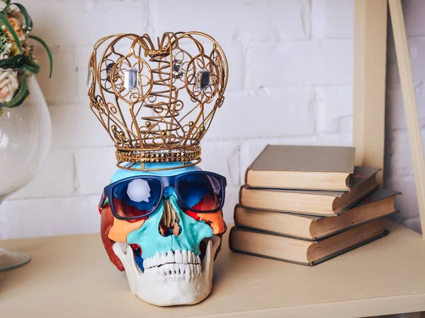 A human skull in sunglasses with a crown and books on the shelf.