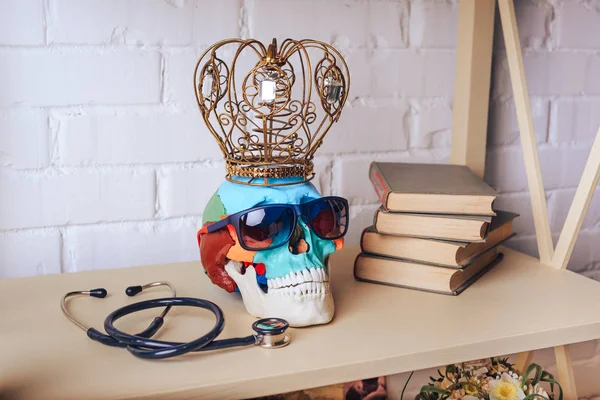 Human skull in sunglasses with crown, stethoscope and books on the shelf.