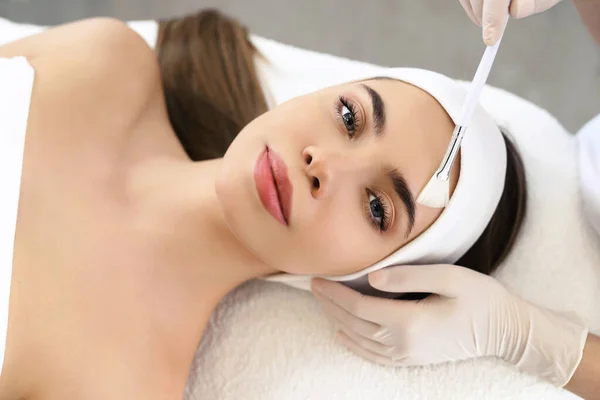 Model in white headband bandage cap lying on couch with closed eyes. Hand of beautician doctor in white glove touching her face with brush. Head and shoulders, healthcare, cosmetology, beauty clinic