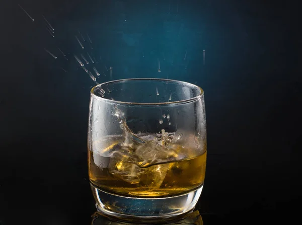 氷が落ちる水しぶきを持つ暗い色の写真に氷とウイスキーのグラス — ストック写真