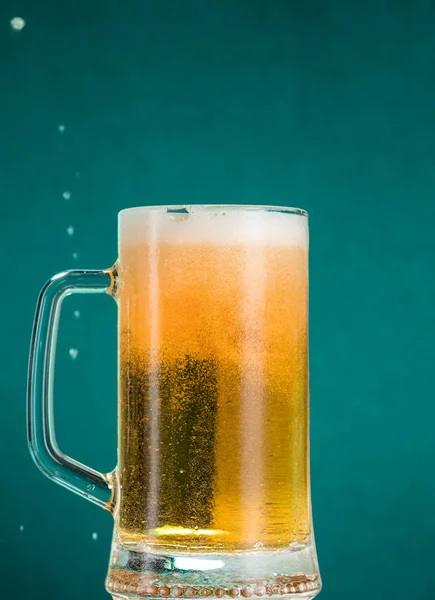 Glass of beer with foam and spray from falling ice on a turquoise green background