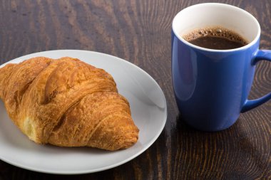 Beyaz tabakta kruvasan ve mor bardakta kahve.