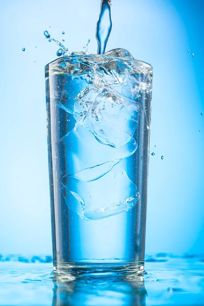 把清澈的水倒入一个杯子里 杯子里装的是蓝色坡度背景的纯冰块 — 图库照片