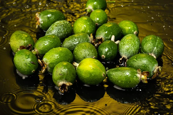 several feijoa fruits with drops and splashes of water on dark glass with gradient background and reflection