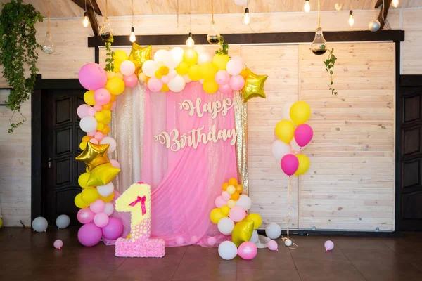Bright decorations to celebrate the first birthday of a child. Pink, yellow and white balloons on a soft pink background. Inscription Happy Birthday.