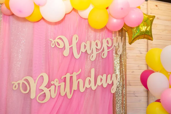 The inscription Happy Birthday made of light wood on a soft pink background. Birthday party decor. Balloons in pink, yellow and white.