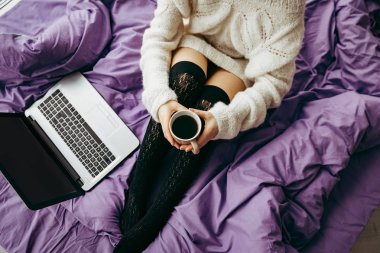 Genç esmer kadın kahve içme ve dizüstü yatağın üstüne oturan beyaz kullanma 