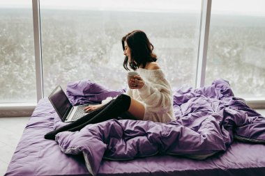 Genç esmer kadın kahve içme ve dizüstü yatağın üstüne oturan beyaz kullanma 