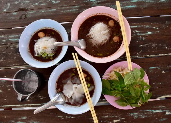 Tayland noodle-domuz eti ve et topu ile Kalınlaşmak çorba. Bir çanak sebze ve fasulye filiz vardır. Paslanmaz cam içleri içerir ve su erişte kase yanında. Yukarıdaki tüm ahşap masada.