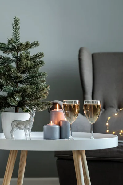 New Year\'s decor: two glasses with champagne, burning candles, a Christmas tree and a decorative statue of a deer on a white table