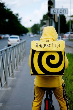 Kazan, Russia - June 07, 2020: Yandex Food - food delivery service. Courier or delivery man with a yellow backpack on a bicycle. Yandex eda. clipart